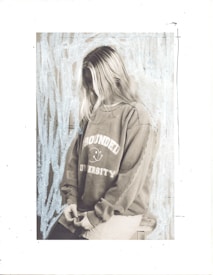 A person with long hair is wearing a sweatshirt with the words 'Grounded University' printed on it. The individual is sitting on a stool or chair with their head slightly bowed and hands clasped together. The background features a textured or artistic pattern, possibly created with light blue strokes.
