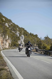Two motorbikes cruising on a cliffside road with waves crashing below under a bright blue sky.