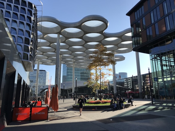 A modern urban plaza with distinctive circular canopies suspended above the open space. The area features contemporary architecture, with glass-fronted buildings on either side and a circular planter with a small tree in the center. People are walking and sitting around the plaza under a clear blue sky.