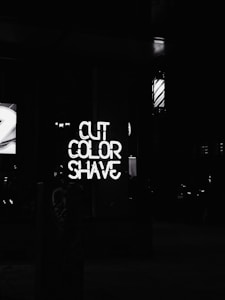 Neon sign displaying the words 'CUT', 'COLOR', 'SHAVE' in a vertical arrangement. The background is dark, possibly indicating nighttime or a dimly lit area. A barber pole with contrasting stripes is visible, hinting at a barbershop or salon setting.