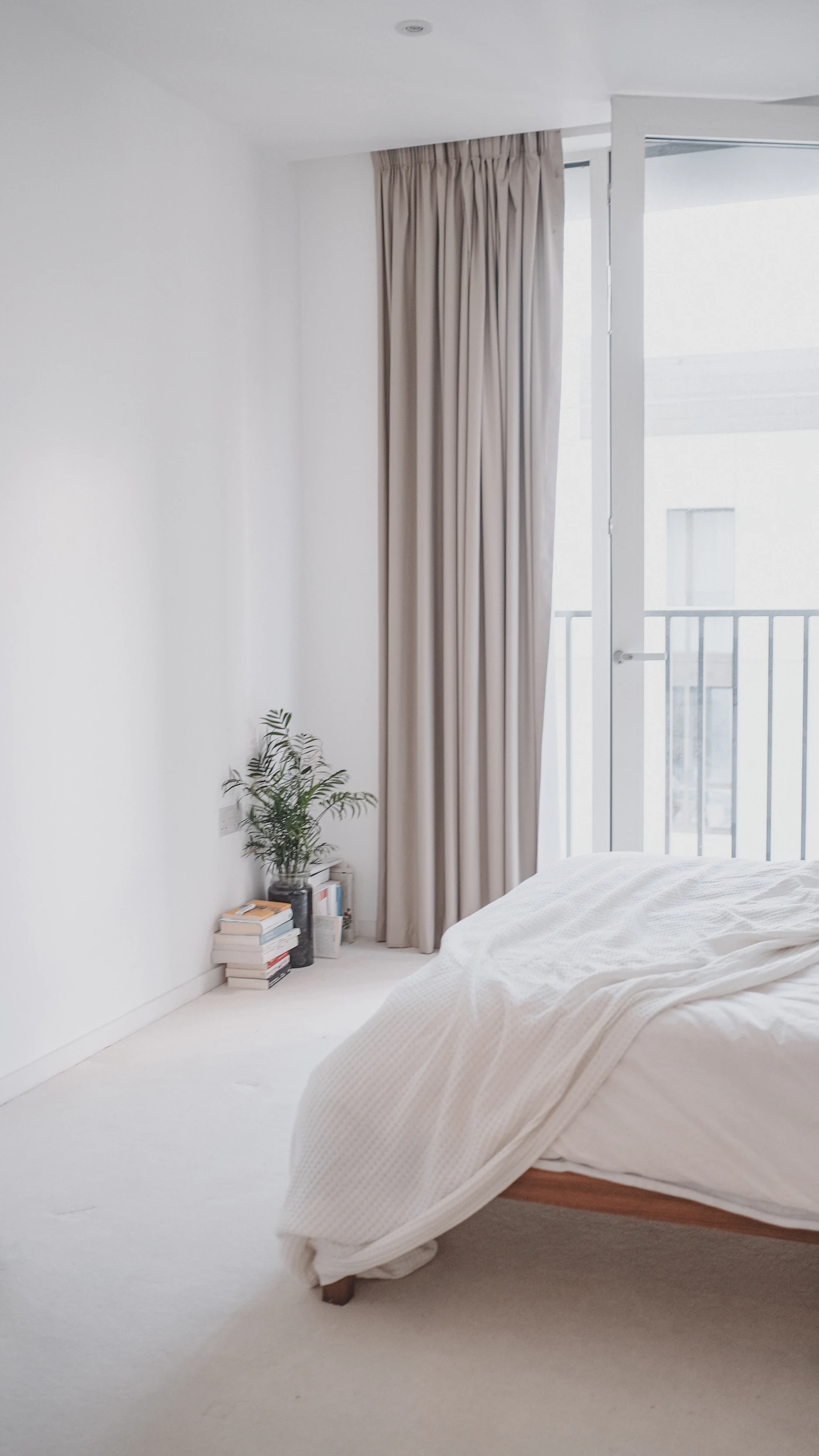 Bright minimalist bedroom with white bedding, a textured throw, full-length beige curtains, a leafy plant, books and a balcony doorway.
