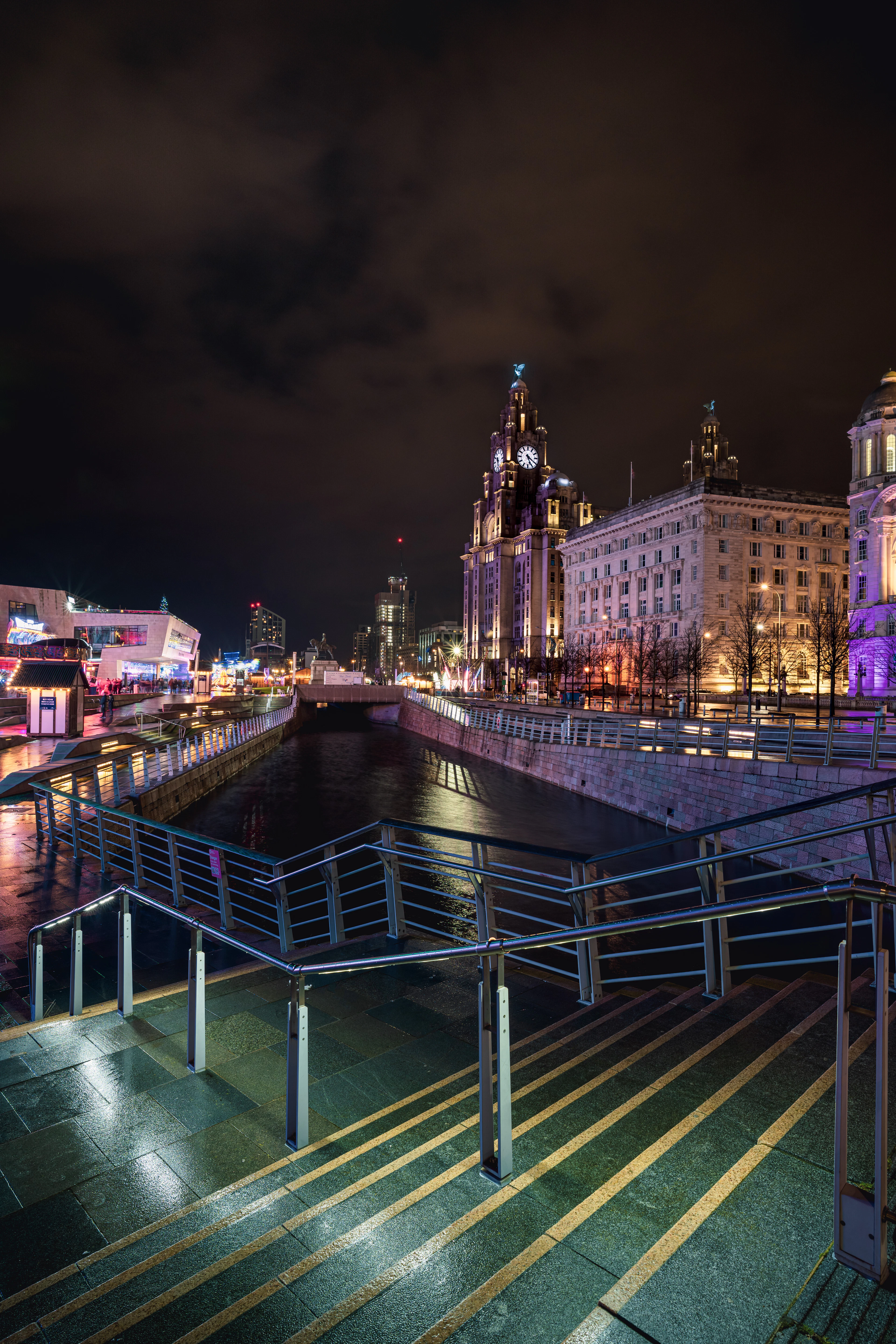 Liverpool's Pier Head at night, the floodlit Royal Liver Building and Cunard Building reflected in the canal link, with wet paving and steel railings in the foreground