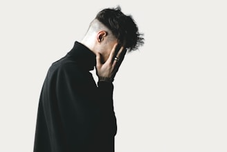 A person with short hair, wearing a dark turtleneck, is facing sideways with their hand covering their face, in a gesture that suggests contemplation or distress. The background is a neutral light color, providing contrast with the dark clothing.