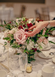 A beautifully arranged floral centerpiece with a variety of roses in shades of pink and red, surrounded by lush greenery and white flowers. A hand with manicured nails and a watch is gently touching the arrangement. The table is elegantly set with glassware and chinaware, creating a sophisticated and romantic atmosphere.
