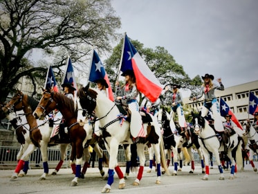 A group of people, dressed in coordinated outfits with cowboy hats, ride on horseback in a parade. They are holding flags with blue and white stars and one distinctive flag with a red, white, and blue pattern, resembling the Texas state flag. The horses are decorated with matching ribbons. The background includes large trees and a few buildings.
