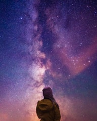 A person in a brown jacket and purple beanie stands gazing up at a vibrant, star-filled night sky featuring the Milky Way. The scene conveys a sense of wonder and awe as the vastness of space is spread across the view with a spectrum of purples, blues, and pinks.