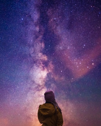 A person in a brown jacket and purple beanie stands gazing up at a vibrant, star-filled night sky featuring the Milky Way. The scene conveys a sense of wonder and awe as the vastness of space is spread across the view with a spectrum of purples, blues, and pinks.