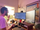A person with a beard, wearing glasses and a white shirt, is working at a desk with a dual-monitor setup, displaying code on the screens. A microphone with a red light is placed on the desk along with a mechanical keyboard and a Rubik’s cube. To the side, there is a guitar hanging on the wall and a framed artwork. Natural light is coming through a window, creating a soft and warm ambiance.