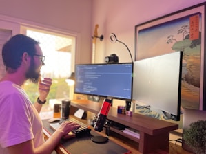 A person with a beard, wearing glasses and a white shirt, is working at a desk with a dual-monitor setup, displaying code on the screens. A microphone with a red light is placed on the desk along with a mechanical keyboard and a Rubik’s cube. To the side, there is a guitar hanging on the wall and a framed artwork. Natural light is coming through a window, creating a soft and warm ambiance.