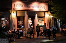 Exterior view of a lively restaurant with a warmly lit entrance. People are gathered around tables enjoying food and drinks, both inside and outside the venue. The sign above the entrance reads 'Tigre Morado'. The ambience is inviting and relaxed, suitable for socializing in the evening.