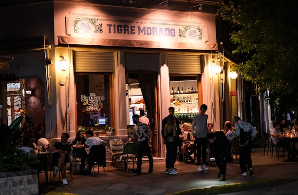 Exterior view of a lively restaurant with a warmly lit entrance. People are gathered around tables enjoying food and drinks, both inside and outside the venue. The sign above the entrance reads 'Tigre Morado'. The ambience is inviting and relaxed, suitable for socializing in the evening.