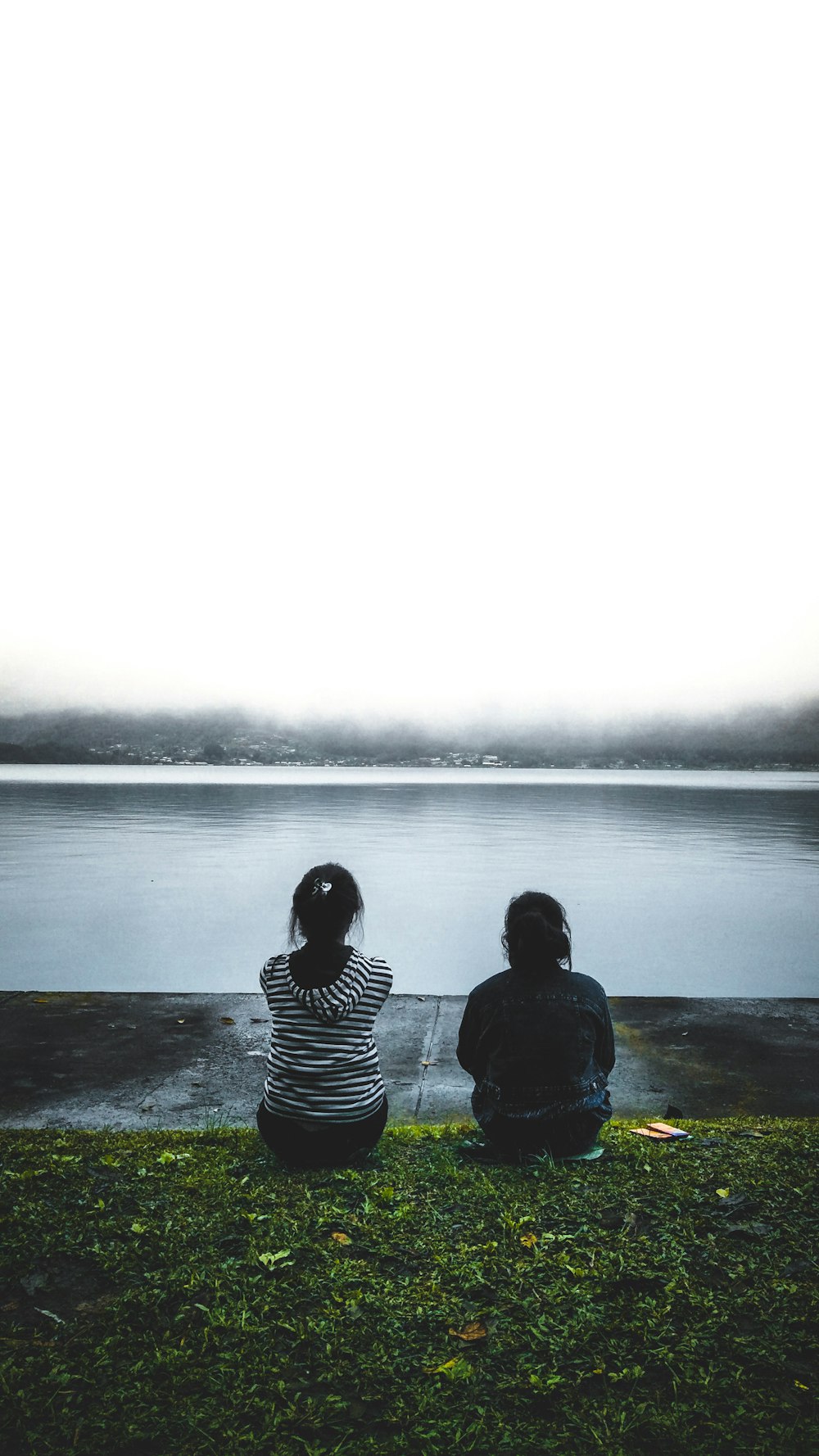 two people sitting on the grass looking out at the water