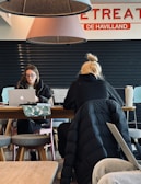 Two individuals are seated at a wooden table, working on laptops. The person on the left is wearing glasses and appears focused on the screen, while the one on the right has blond hair tied in a bun and is depicted from behind. Overhead, two large cone-shaped lamps hang from the ceiling with parts of a sign saying 'RETREAT' and 'DE HAVILLAND' visible. A patterned bag and a large drink cup are on the table. The setting looks like a cafe or shared workspace with a few stools around.