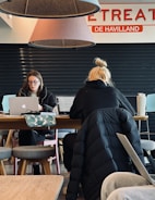 Two individuals are seated at a wooden table, working on laptops. The person on the left is wearing glasses and appears focused on the screen, while the one on the right has blond hair tied in a bun and is depicted from behind. Overhead, two large cone-shaped lamps hang from the ceiling with parts of a sign saying 'RETREAT' and 'DE HAVILLAND' visible. A patterned bag and a large drink cup are on the table. The setting looks like a cafe or shared workspace with a few stools around.