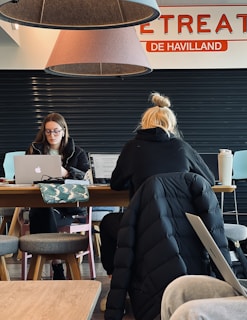 Two individuals are seated at a wooden table, working on laptops. The person on the left is wearing glasses and appears focused on the screen, while the one on the right has blond hair tied in a bun and is depicted from behind. Overhead, two large cone-shaped lamps hang from the ceiling with parts of a sign saying 'RETREAT' and 'DE HAVILLAND' visible. A patterned bag and a large drink cup are on the table. The setting looks like a cafe or shared workspace with a few stools around.