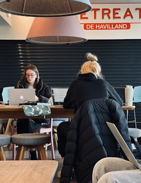 Two individuals are seated at a wooden table, working on laptops. The person on the left is wearing glasses and appears focused on the screen, while the one on the right has blond hair tied in a bun and is depicted from behind. Overhead, two large cone-shaped lamps hang from the ceiling with parts of a sign saying 'RETREAT' and 'DE HAVILLAND' visible. A patterned bag and a large drink cup are on the table. The setting looks like a cafe or shared workspace with a few stools around.