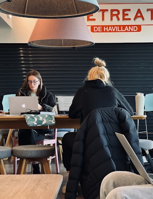 Two individuals are seated at a wooden table, working on laptops. The person on the left is wearing glasses and appears focused on the screen, while the one on the right has blond hair tied in a bun and is depicted from behind. Overhead, two large cone-shaped lamps hang from the ceiling with parts of a sign saying 'RETREAT' and 'DE HAVILLAND' visible. A patterned bag and a large drink cup are on the table. The setting looks like a cafe or shared workspace with a few stools around.