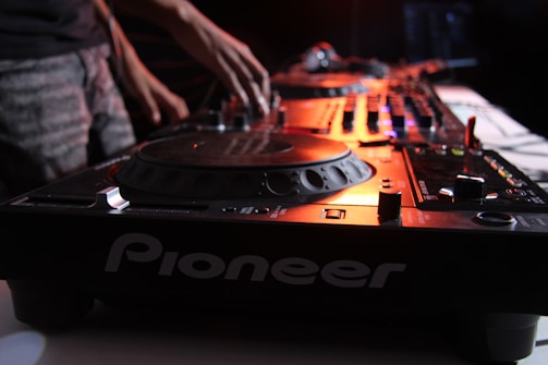 A close-up of a DJ's hand adjusting the controls on a Pioneer DJ mixer with a turntable. The lighting is dim and moody, highlighting the equipment with a warm, orange glow.