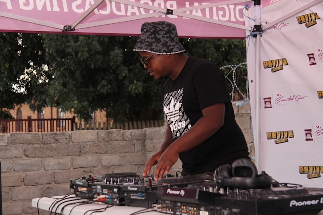 A person wearing a bucket hat and sunglasses is DJing at an outdoor event. They are using a Pioneer DJ controller on a table with various cables and equipment. The background features a brick wall, trees, and a canopy with branding and logos.