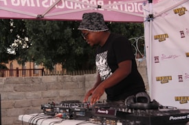 A person wearing a bucket hat and sunglasses is DJing at an outdoor event. They are using a Pioneer DJ controller on a table with various cables and equipment. The background features a brick wall, trees, and a canopy with branding and logos.