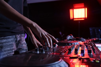 A person is using a DJ mixer with various knobs and sliders under low lighting conditions. The scene is illuminated by a red light, creating a club-like atmosphere. A pair of headphones is placed on the mixer, ready for use.