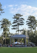 People are engaging in a group exercise session outside, in front of a large pavilion surrounded by tall trees. The sky is partially cloudy, and the area is lush with greenery.