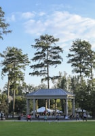People are engaging in a group exercise session outside, in front of a large pavilion surrounded by tall trees. The sky is partially cloudy, and the area is lush with greenery.