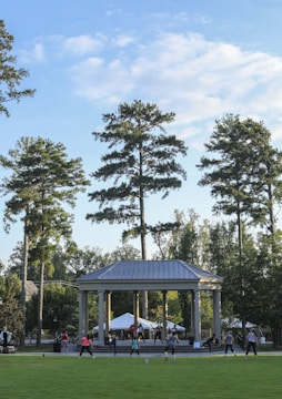 People are engaging in a group exercise session outside, in front of a large pavilion surrounded by tall trees. The sky is partially cloudy, and the area is lush with greenery.