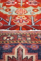 An intricate and colorful textile featuring detailed geometric patterns and motifs. The upper section displays a red background with symmetric shapes in various colors, including green, yellow, and pink, resembling floral and abstract forms. The lower section has a darker palette with a pattern that includes a mix of dark blue, red, and cream, bordered with a distinct geometric design.