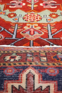 An intricate and colorful textile featuring detailed geometric patterns and motifs. The upper section displays a red background with symmetric shapes in various colors, including green, yellow, and pink, resembling floral and abstract forms. The lower section has a darker palette with a pattern that includes a mix of dark blue, red, and cream, bordered with a distinct geometric design.