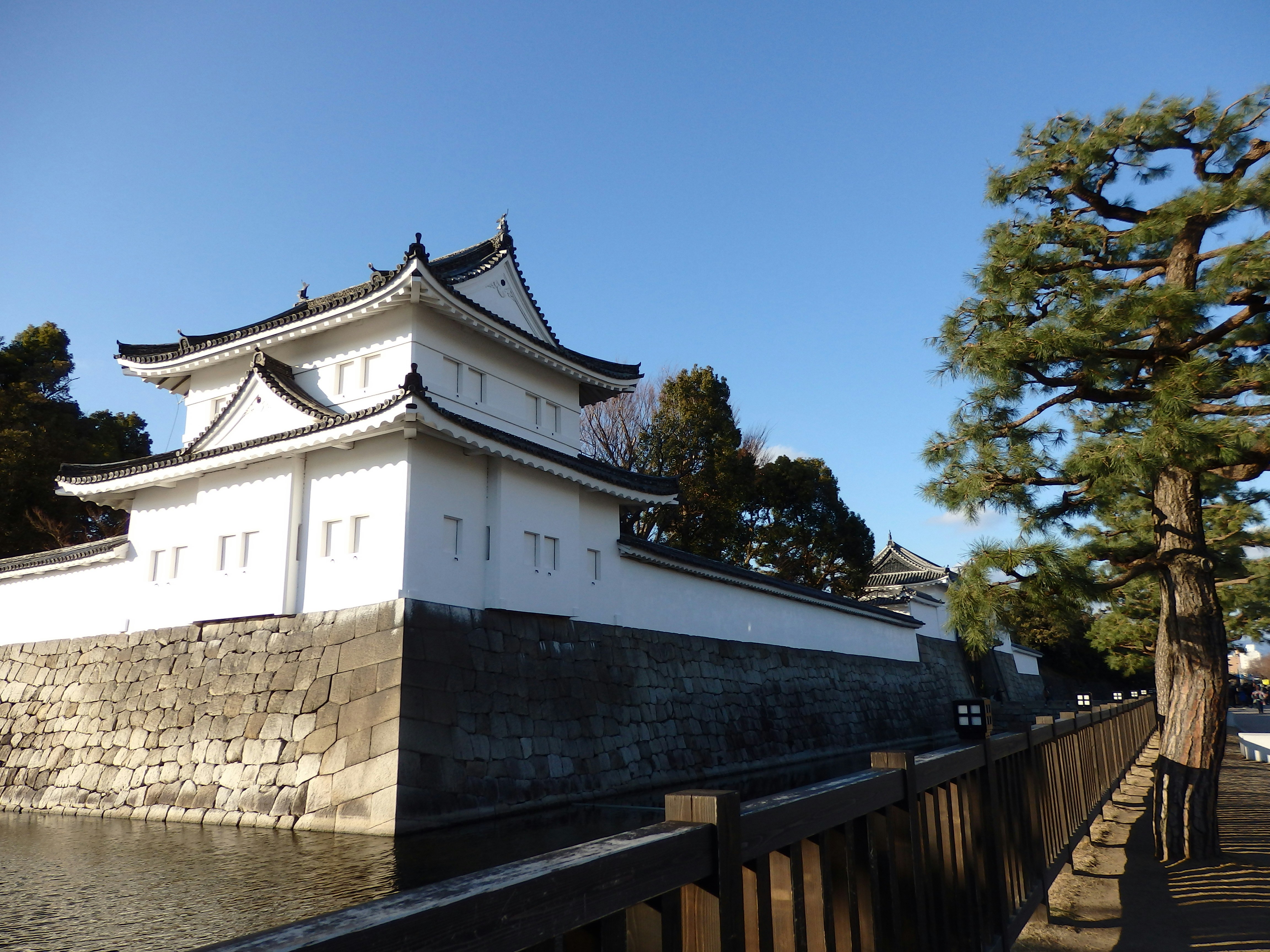 A photograph of a white-walled Japanese castle perched on a stone base beside a calm moat, with a wooden railing and tall trees lining the path.