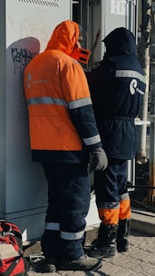 Two workers wearing protective clothing are standing in front of an electrical panel. They are dressed in heavy-duty jackets with hoods, one in orange and the other in dark blue. The surroundings suggest an industrial or outdoor setting, with a backpack lying on the ground nearby.