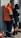 Two workers wearing protective clothing are standing in front of an electrical panel. They are dressed in heavy-duty jackets with hoods, one in orange and the other in dark blue. The surroundings suggest an industrial or outdoor setting, with a backpack lying on the ground nearby.