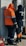 Two workers wearing protective clothing are standing in front of an electrical panel. They are dressed in heavy-duty jackets with hoods, one in orange and the other in dark blue. The surroundings suggest an industrial or outdoor setting, with a backpack lying on the ground nearby.