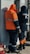 Two workers wearing protective clothing are standing in front of an electrical panel. They are dressed in heavy-duty jackets with hoods, one in orange and the other in dark blue. The surroundings suggest an industrial or outdoor setting, with a backpack lying on the ground nearby.
