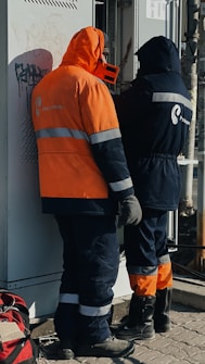 Two workers wearing protective clothing are standing in front of an electrical panel. They are dressed in heavy-duty jackets with hoods, one in orange and the other in dark blue. The surroundings suggest an industrial or outdoor setting, with a backpack lying on the ground nearby.
