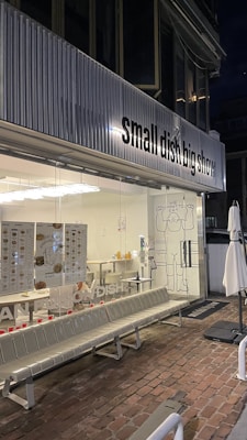 A modern restaurant exterior with a clear glass storefront displaying a sign that reads 'small dish big show'. Metallic seating is arranged outside on a brick pathway. The interior is well-lit with visible menus and a playful cartoon figure drawing on the glass door.