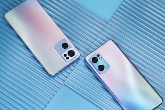 Two smartphones are placed on a textured blue surface. The phones have a sleek design with a gradient color scheme and prominent camera modules on the back.