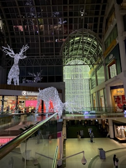 A shopping mall with large, illuminated reindeer decorations made of white lights. The interior features a high glass ceiling and multiple levels with stores such as Lululemon and Indigo. The lighting creates a festive ambiance, and there are few people visible on the lower level.