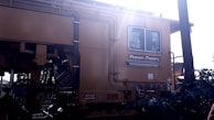 A large yellow industrial machine with various pipes and cables is featured prominently. The brand 'Plasser & Theurer' is visible on the side. The machine has a window, and there are signs indicating safety precautions and danger areas. Sunlight is streaming from the upper right corner, creating a bright glare.