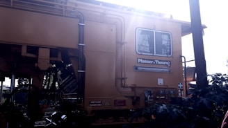 A large yellow industrial machine with various pipes and cables is featured prominently. The brand 'Plasser & Theurer' is visible on the side. The machine has a window, and there are signs indicating safety precautions and danger areas. Sunlight is streaming from the upper right corner, creating a bright glare.