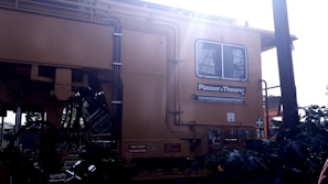 A large yellow industrial machine with various pipes and cables is featured prominently. The brand 'Plasser & Theurer' is visible on the side. The machine has a window, and there are signs indicating safety precautions and danger areas. Sunlight is streaming from the upper right corner, creating a bright glare.