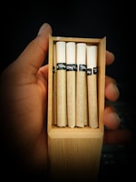 A hand holding an open wooden box containing five rolled cigarettes. The cigarettes have white tips and brown paper, with black bands featuring text.
