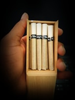 A hand holding an open wooden box containing five rolled cigarettes. The cigarettes have white tips and brown paper, with black bands featuring text.