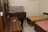 A small room featuring two single beds with patterned bedspreads, a wooden desk with a remote control on it, a swivel chair, and a wall-mounted television. The room has a kitchenette area with a marble countertop, a sink, and some wooden cabinets.