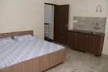 A simple room with a double bed and a small kitchenette. The bed has a geometric-patterned bedspread with two pillows. The kitchenette includes a sink and a cabinet with a light marble backsplash and countertop. The floors are tiled, and there is a wood-paneled door leading to an adjacent area.