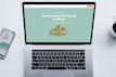 A laptop with a financial website open is centered on a white table. The screen displays a mint green background with text related to commission-free stock trading, accompanied by a gold object resembling a bridge. To the left, a smartphone displays a financial app with a graph and monetary figure. A cup of coffee sits on the right side, adding a casual work environment atmosphere.