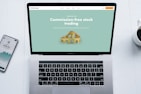 A laptop with a financial website open is centered on a white table. The screen displays a mint green background with text related to commission-free stock trading, accompanied by a gold object resembling a bridge. To the left, a smartphone displays a financial app with a graph and monetary figure. A cup of coffee sits on the right side, adding a casual work environment atmosphere.