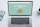 A laptop with a financial website open is centered on a white table. The screen displays a mint green background with text related to commission-free stock trading, accompanied by a gold object resembling a bridge. To the left, a smartphone displays a financial app with a graph and monetary figure. A cup of coffee sits on the right side, adding a casual work environment atmosphere.