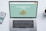 A laptop with a financial website open is centered on a white table. The screen displays a mint green background with text related to commission-free stock trading, accompanied by a gold object resembling a bridge. To the left, a smartphone displays a financial app with a graph and monetary figure. A cup of coffee sits on the right side, adding a casual work environment atmosphere.